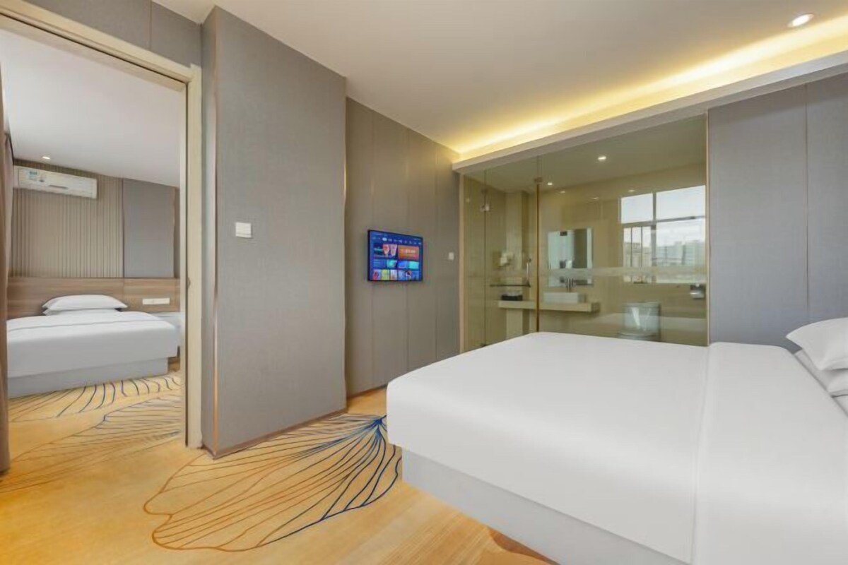 A modern hotel room with a large bed, a bedside table, a TV, and a glass partition separating the room from the bathroom.