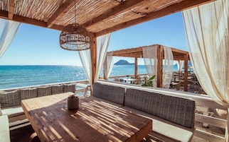 Un pontile in legno con tavolo e sedute imbottite, con vista su una spiaggia e il mare.