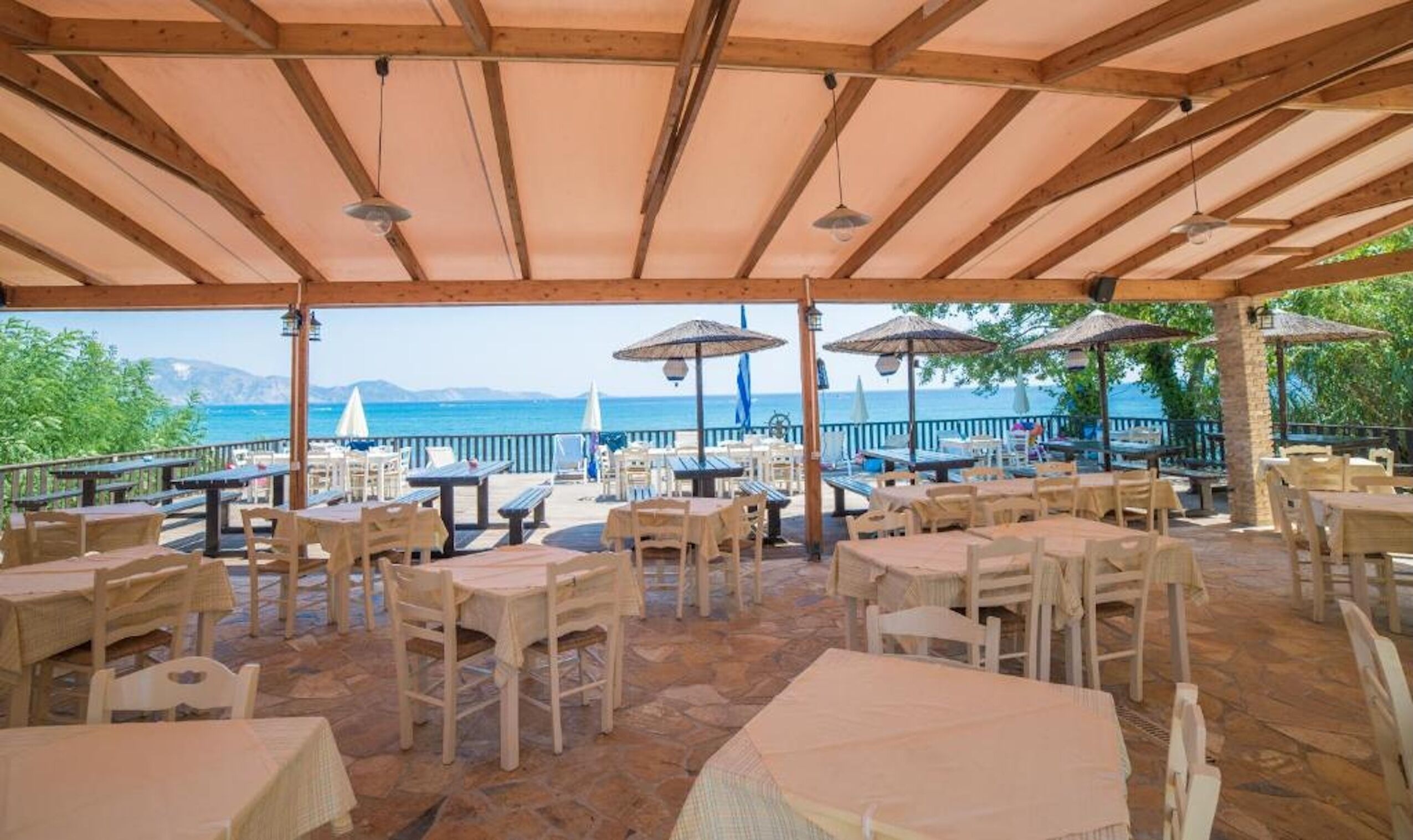 An outdoor patio with tables and chairs, a wooden ceiling, and a view of the sea.