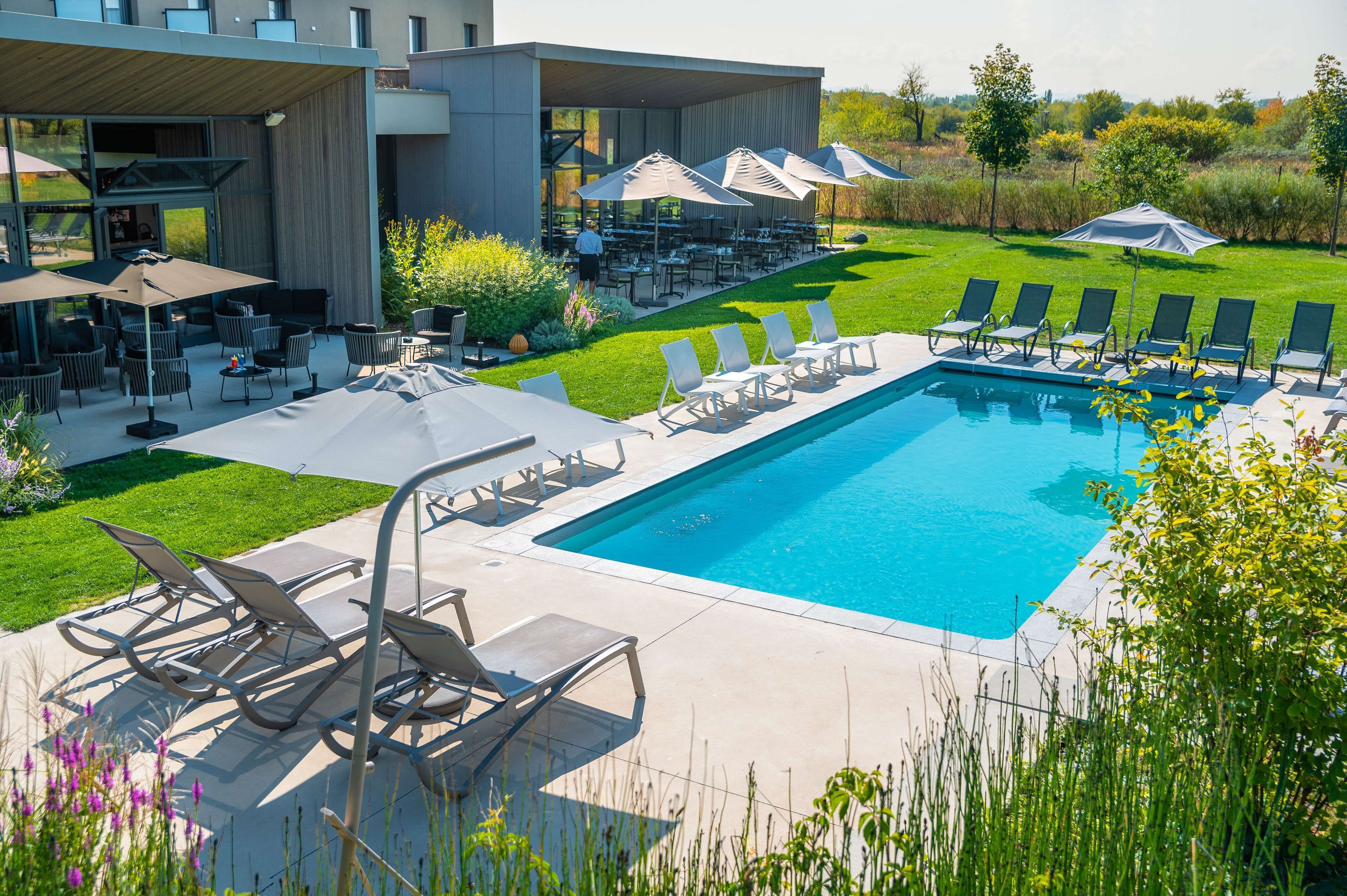 Un área de piscina exterior moderna con sillones de descanso y sombrillas, rodeada por un césped cuidado y un edificio con grandes ventanales.
