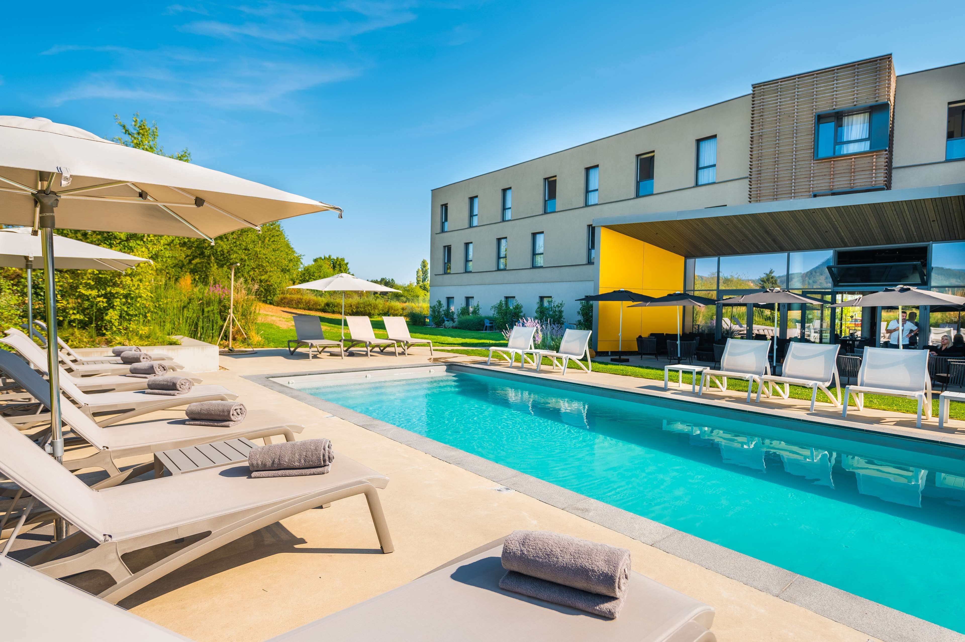 Un área de piscina moderna con sillones y sombrillas, una piscina azul cristalina y un edificio con ventanas amplias y un muro amarillo de acento.