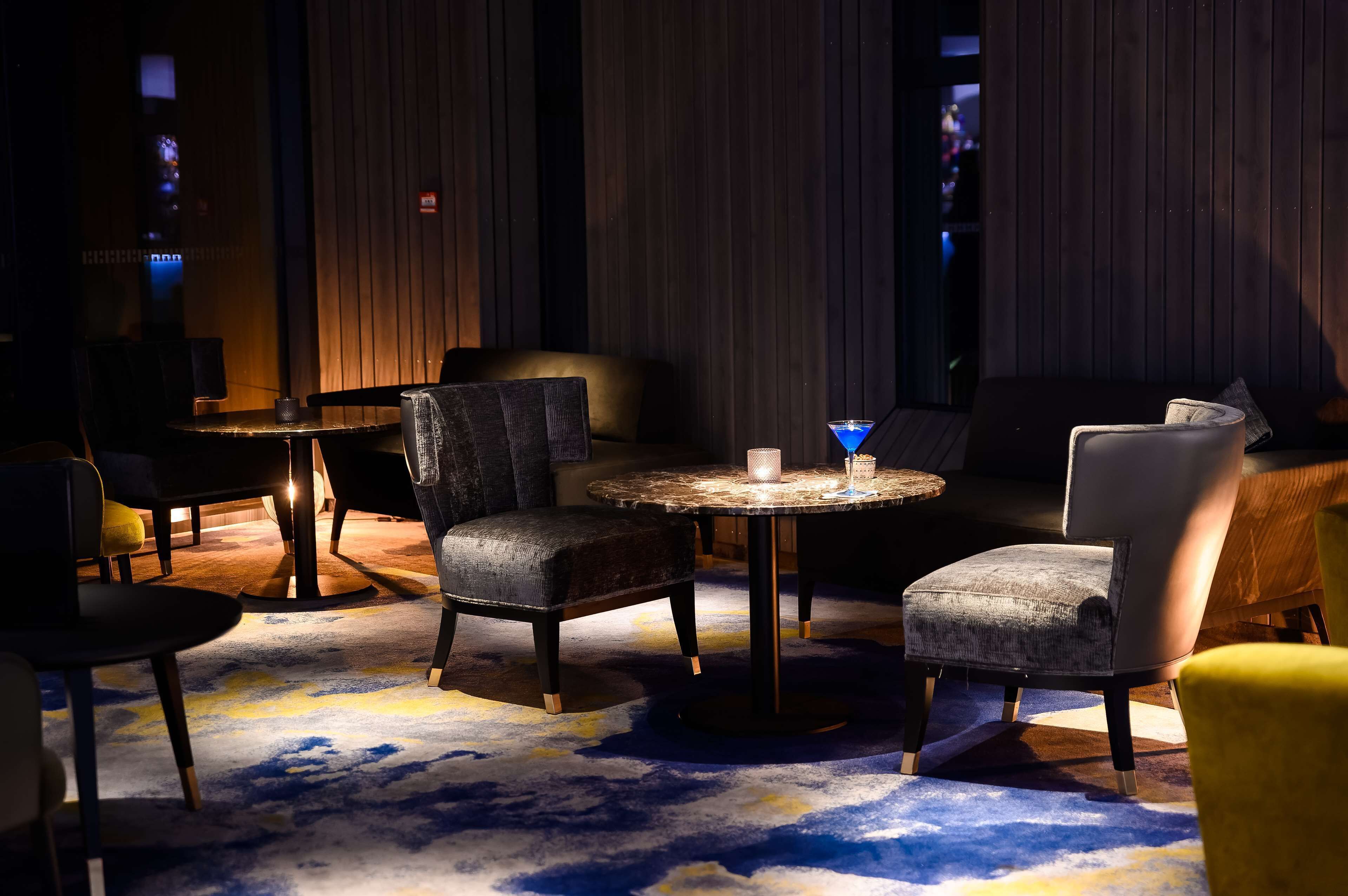 A dimly lit lounge area with round tables and chairs, a patterned carpet, and wooden panel walls.