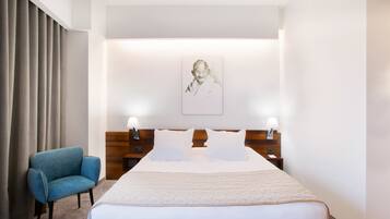 Quarto de hotel com uma cama grande, duas luminárias de cabeceira, cabeceira com um retrato, uma poltrona azul e uma janela com cortinas.