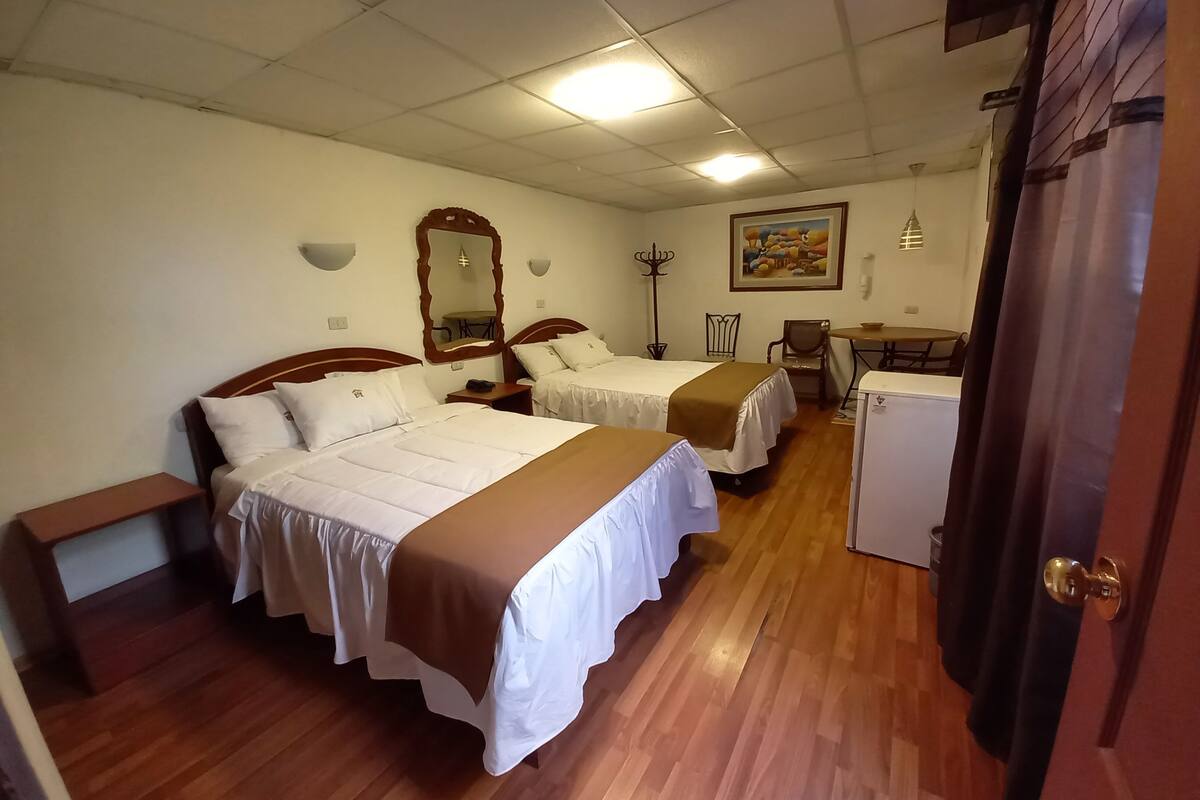 Ett hotellrum med två sängar, ett skrivbord, en stol, ett litet bord, ett kylskåp och en dörr.