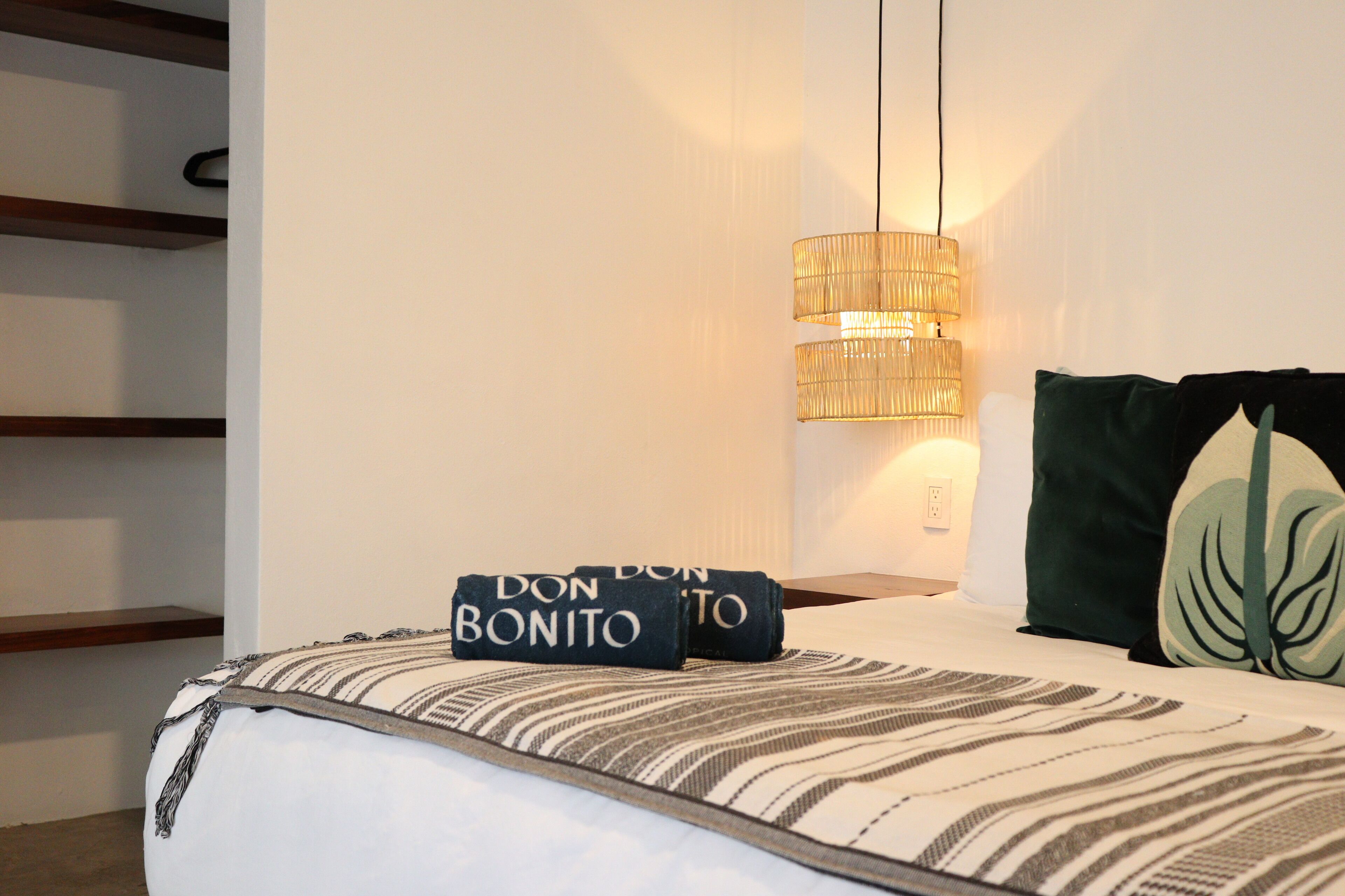 Una cama bien hecha con una colcha a rayas y almohadas, una lámpara colgante y un estante con estructura de madera.