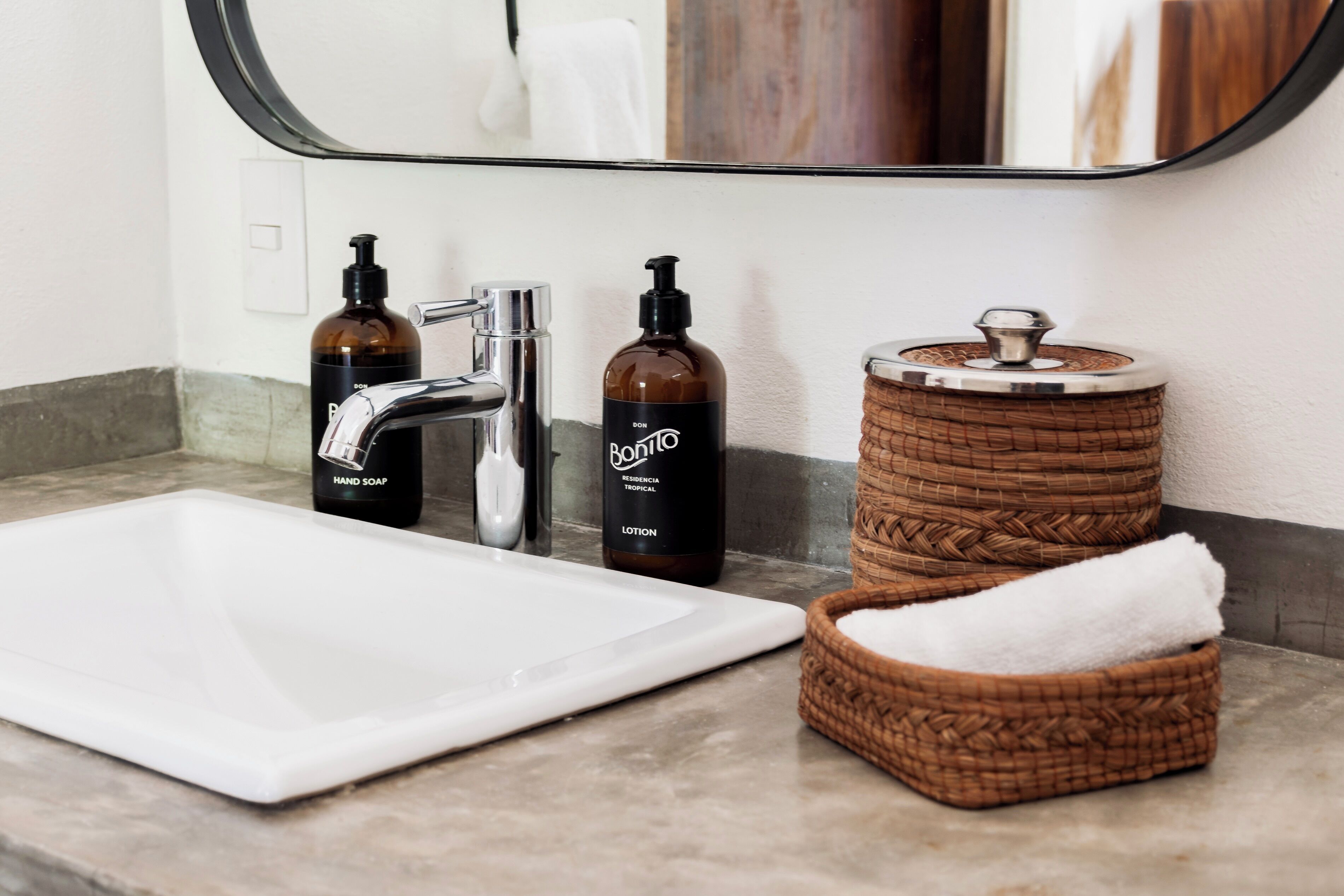 A bathroom with a white sink, two bottles of lotion, a soap dispenser, and a woven basket with a towel.