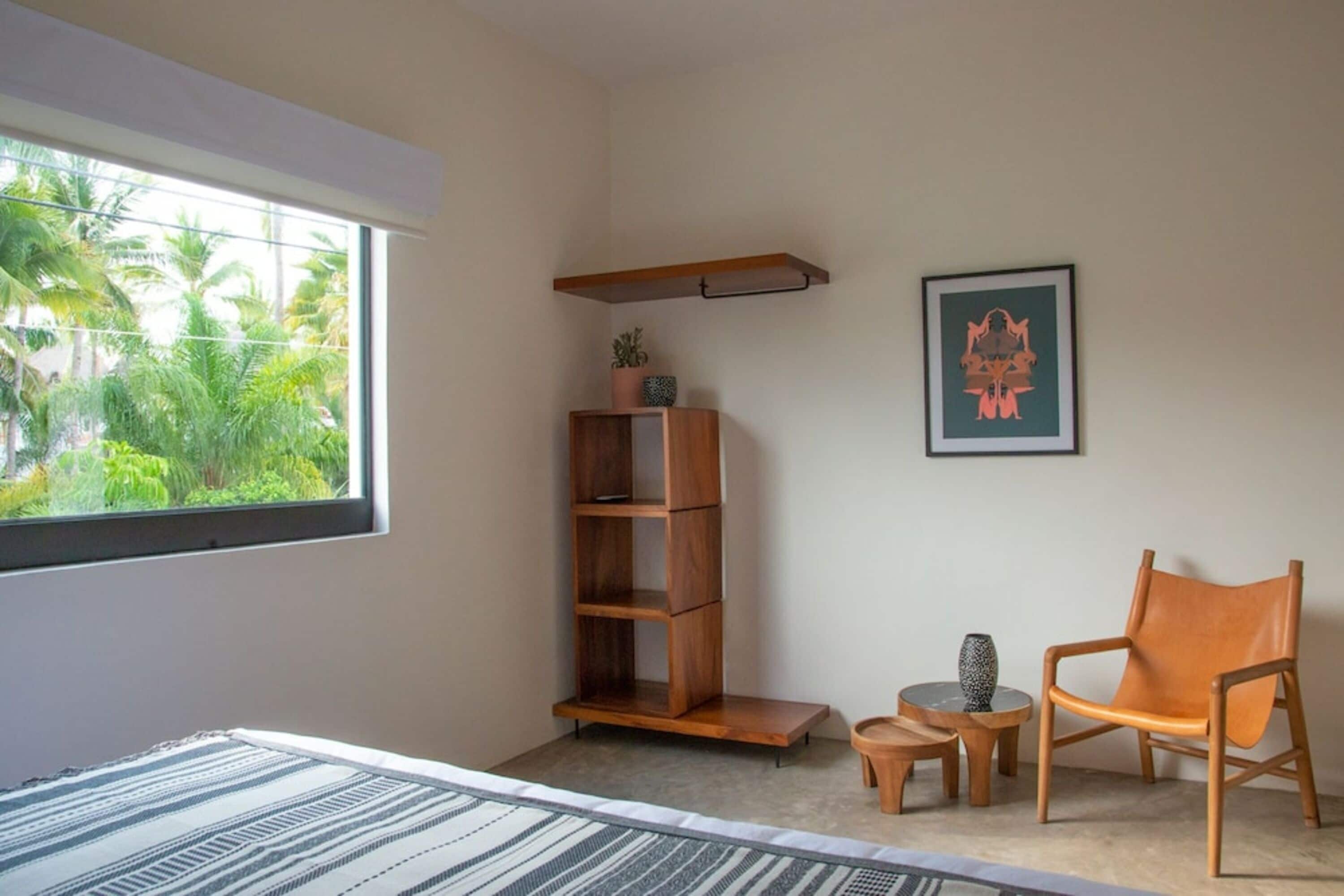 A room with a large window, a wooden bookshelf, a framed artwork, a leather chair, and a small round table with a vase.
