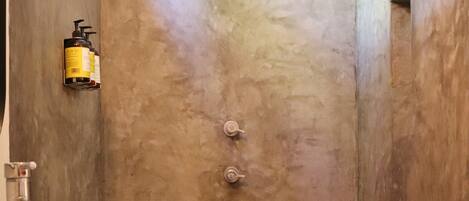 Un baño con un lavamanos grande y blanco, una ducha con una única perilla y una botella de champú en la pared.