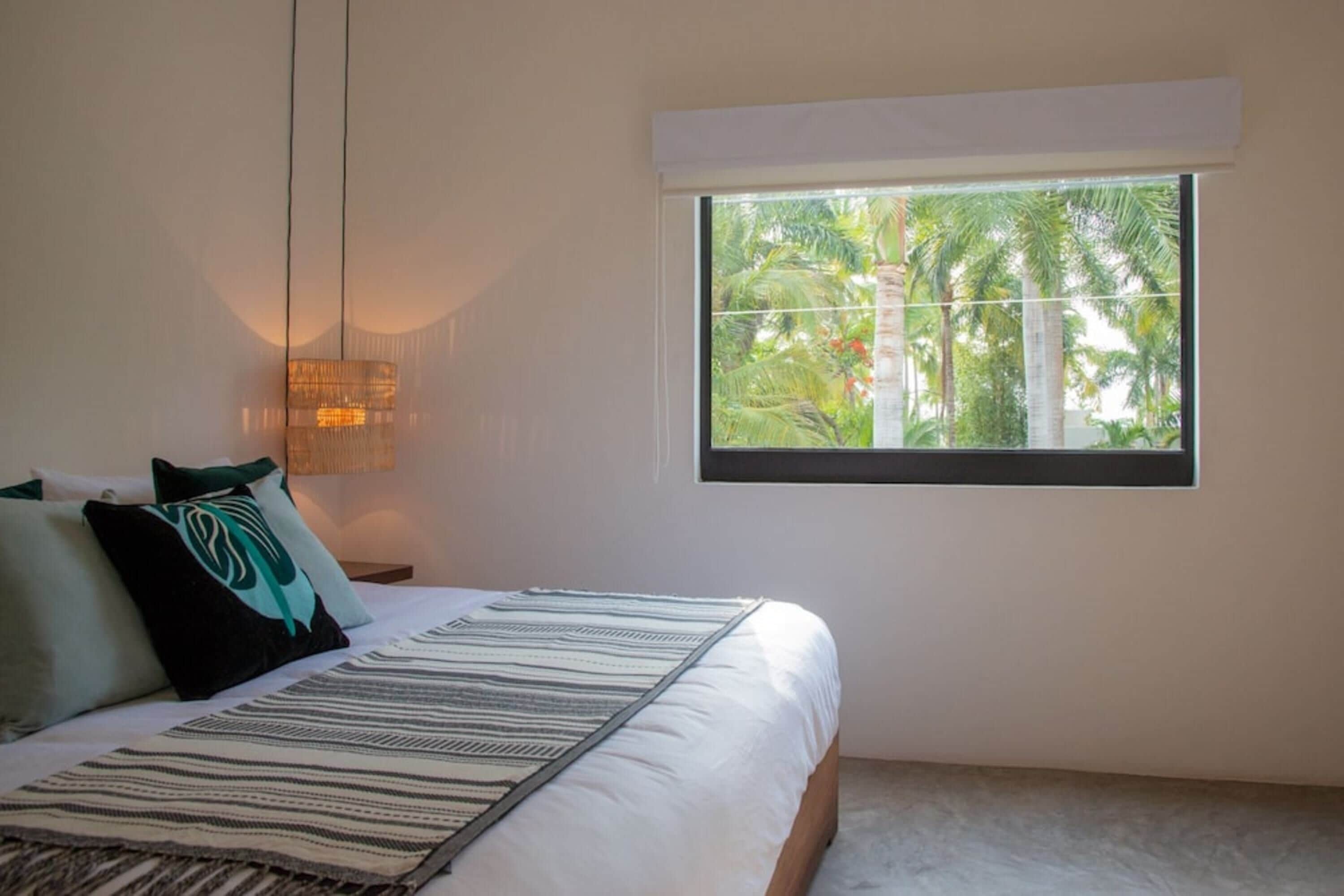 Un dormitorio con cama, almohadas, una ventana con vista a palmeras y una lámpara de pared.