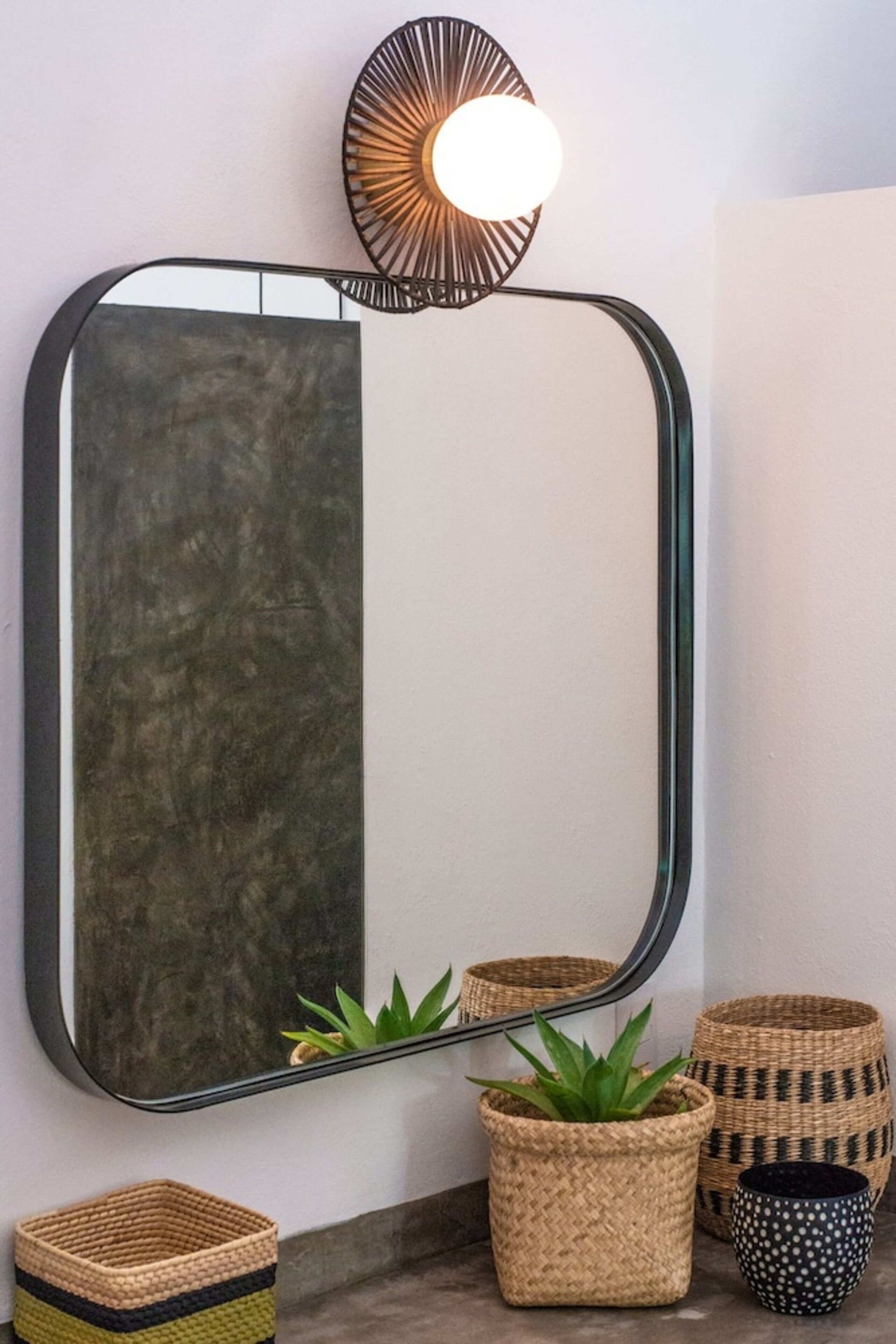 Un miroir avec un cadre noir, un luminaire fixé au mur et trois paniers tressés de différentes tailles et de différents motifs.