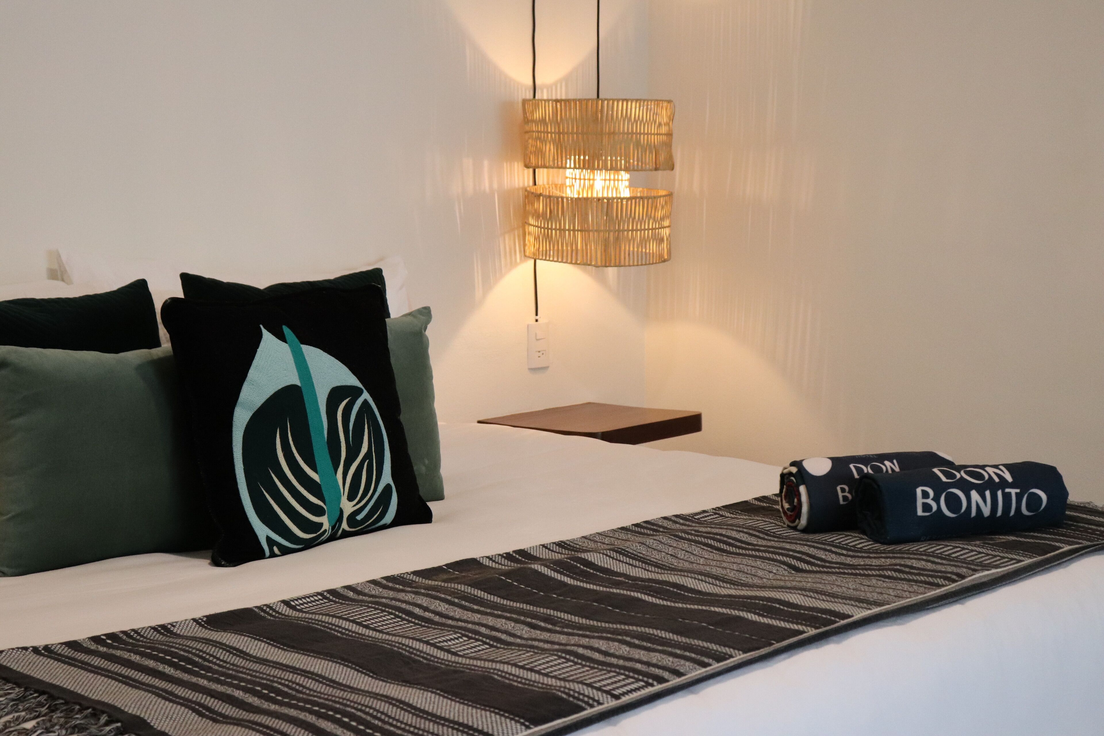 Habitación de hotel ordenada con cama, almohadas, una mesita de noche, lámpara y una toalla enrollada con texto.