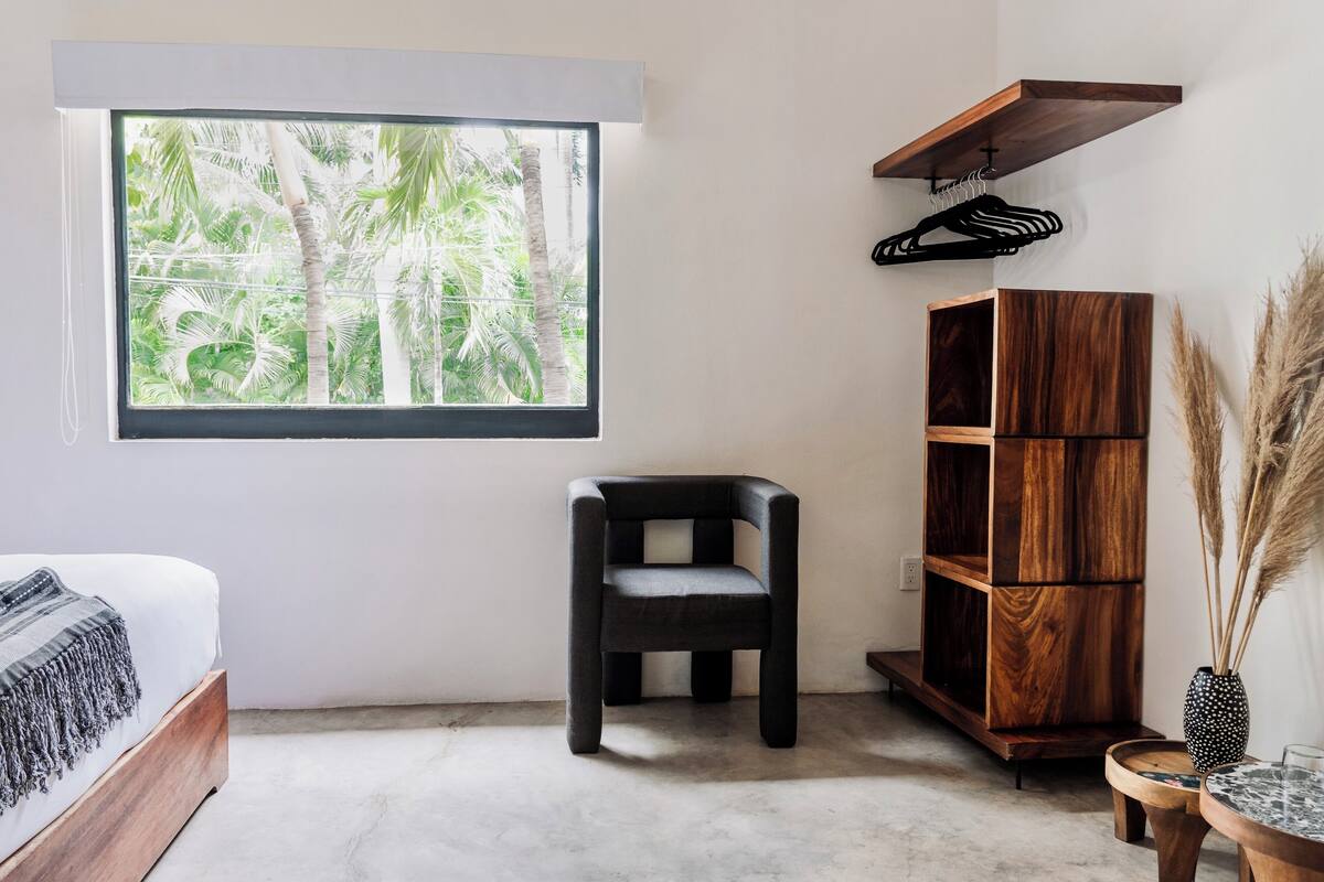Une chambre avec un lit, une fenêtre donnant sur des palmiers, une chaise noire, une bibliothèque en bois et une petite table sur laquelle est posé un vase rempli de plumes d’herbe des pampas.
