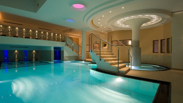A large indoor swimming pool with a central staircase and a modern ceiling design. - Dolce Avita Hotel Spa & Resort (Andalo)