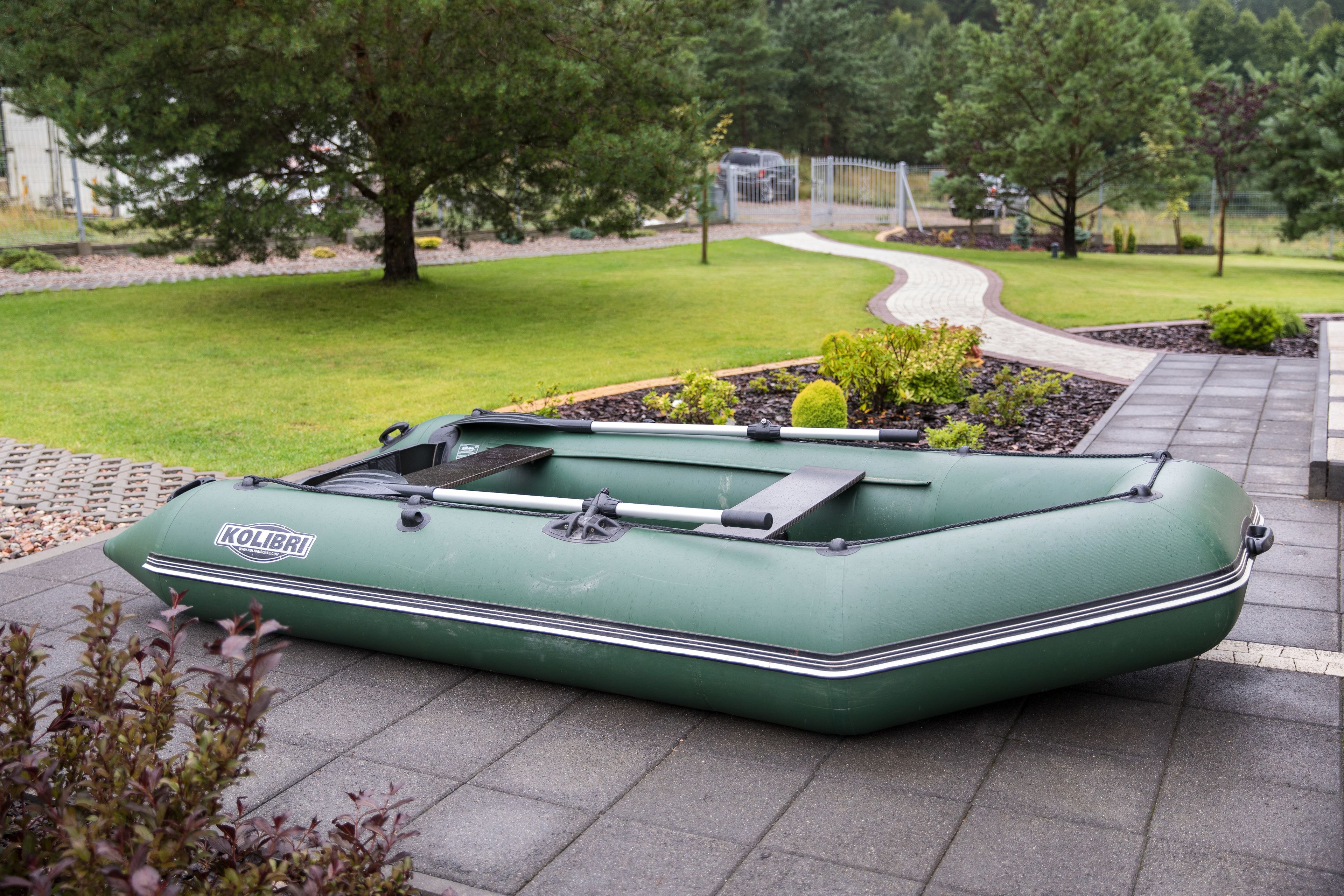 A green inflatable boat with the brand name 'KOLIBRI' on it, parked on a paved area with a grassy lawn and trees in the background.