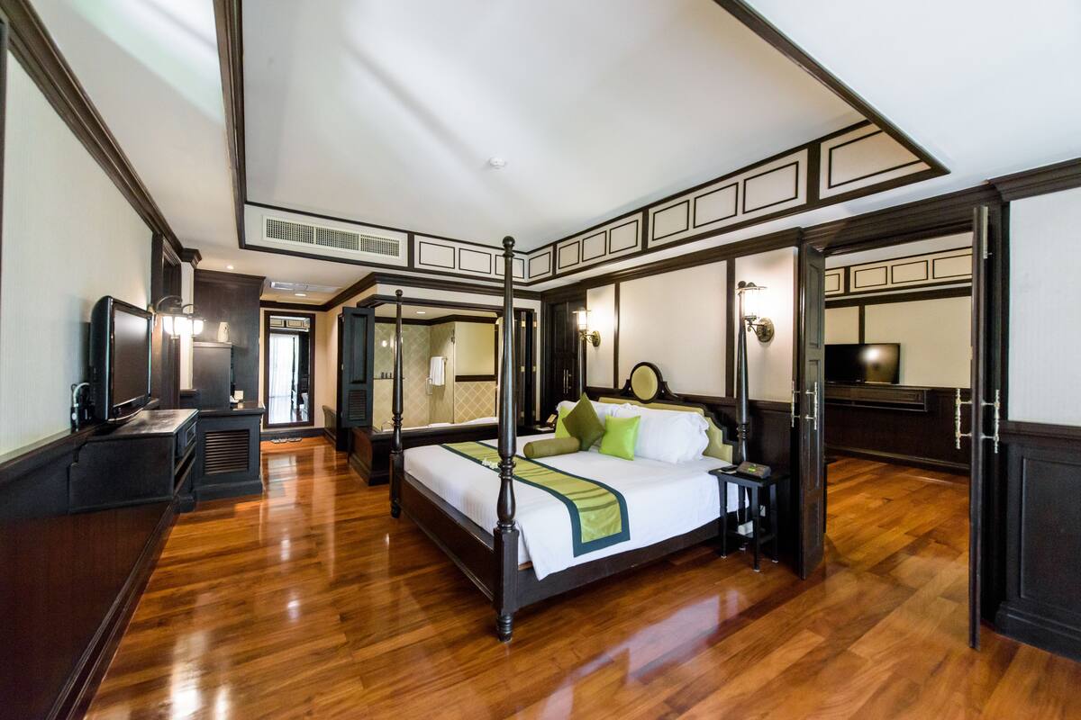 A four-poster bed with a green throw, a television, and a desk lamp in a hotel room with wooden floors and dark trim.
