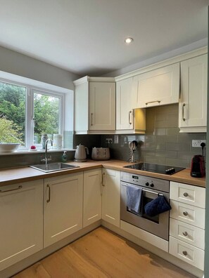 A kitchen with white cabinets, a window with a view, a sink, a stove, and a countertop with various appliances. - The Nook County Dublin (Shankill)
