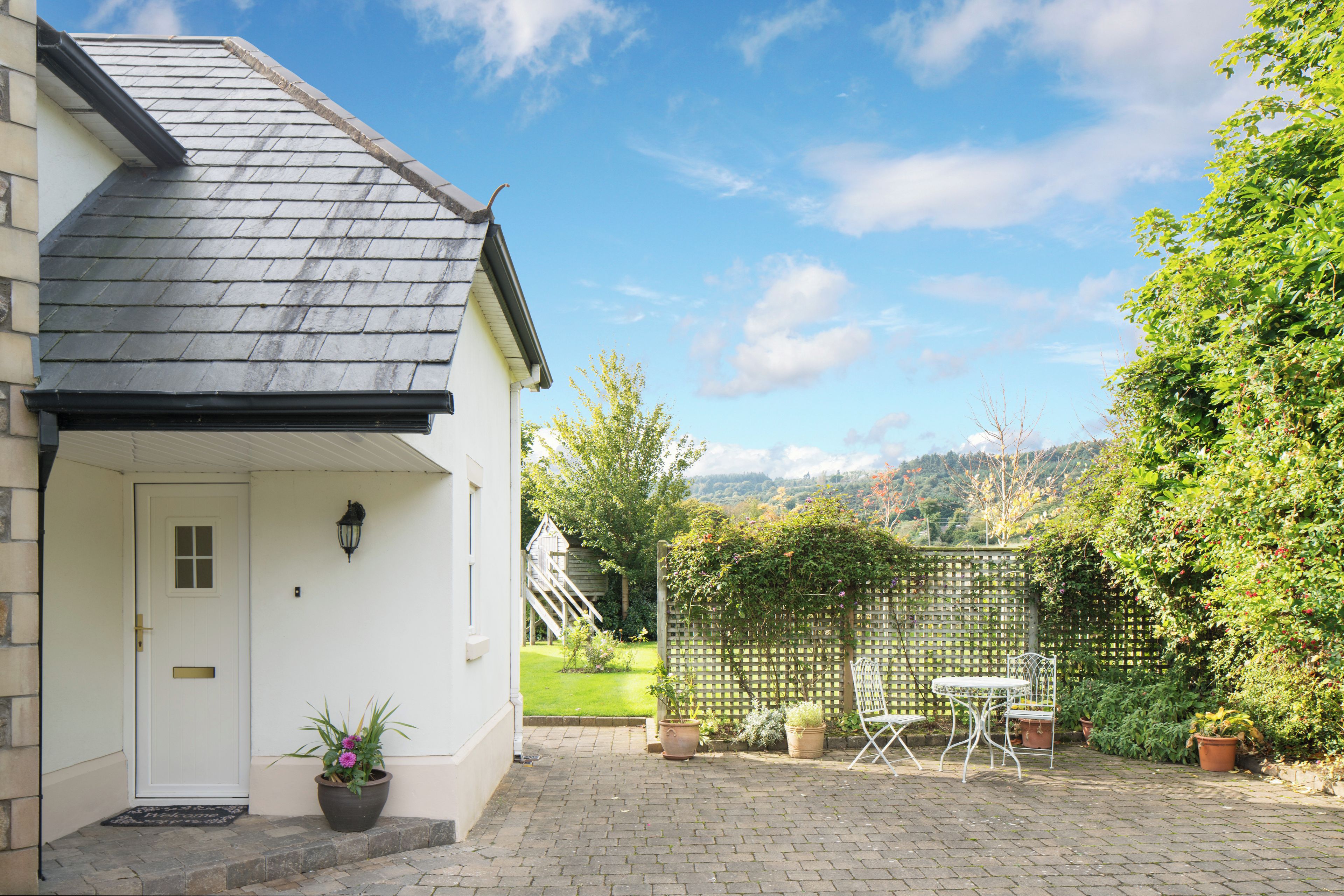 A house with a white door, a small porch, a potted plant, a wooden fence, and a view of a green landscape.