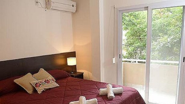 Una cama bien hecha con una colcha roja y dos toallas enrolladas, una mesita de noche con una lámpara, y un ventanal corredero que da a un balcón exterior con vegetación.