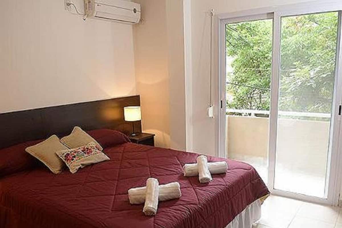 A neatly made bed with a red bedspread and two rolled towels, a nightstand with a lamp, and a sliding glass door leading to an outdoor balcony with greenery.