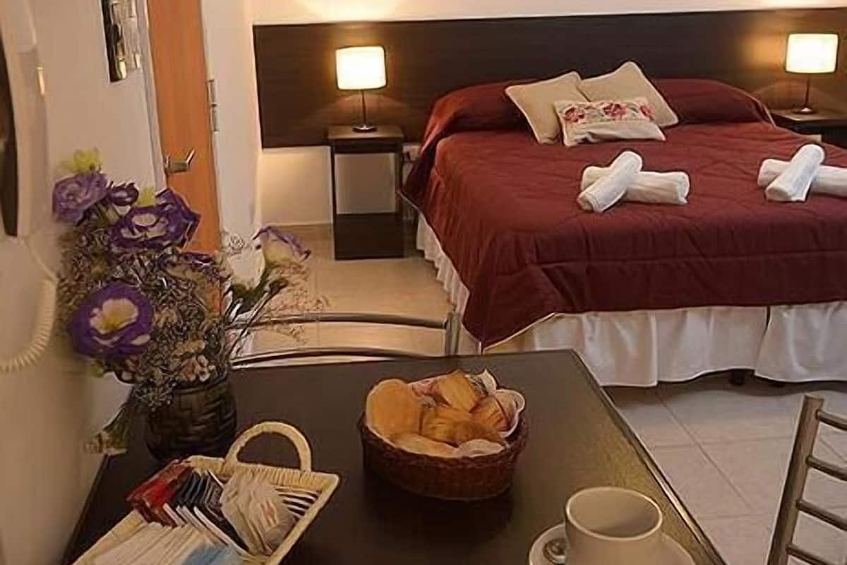 A hotel room with a bed, bedside lamps, a table with a basket of bread, a vase with flowers, and a telephone.