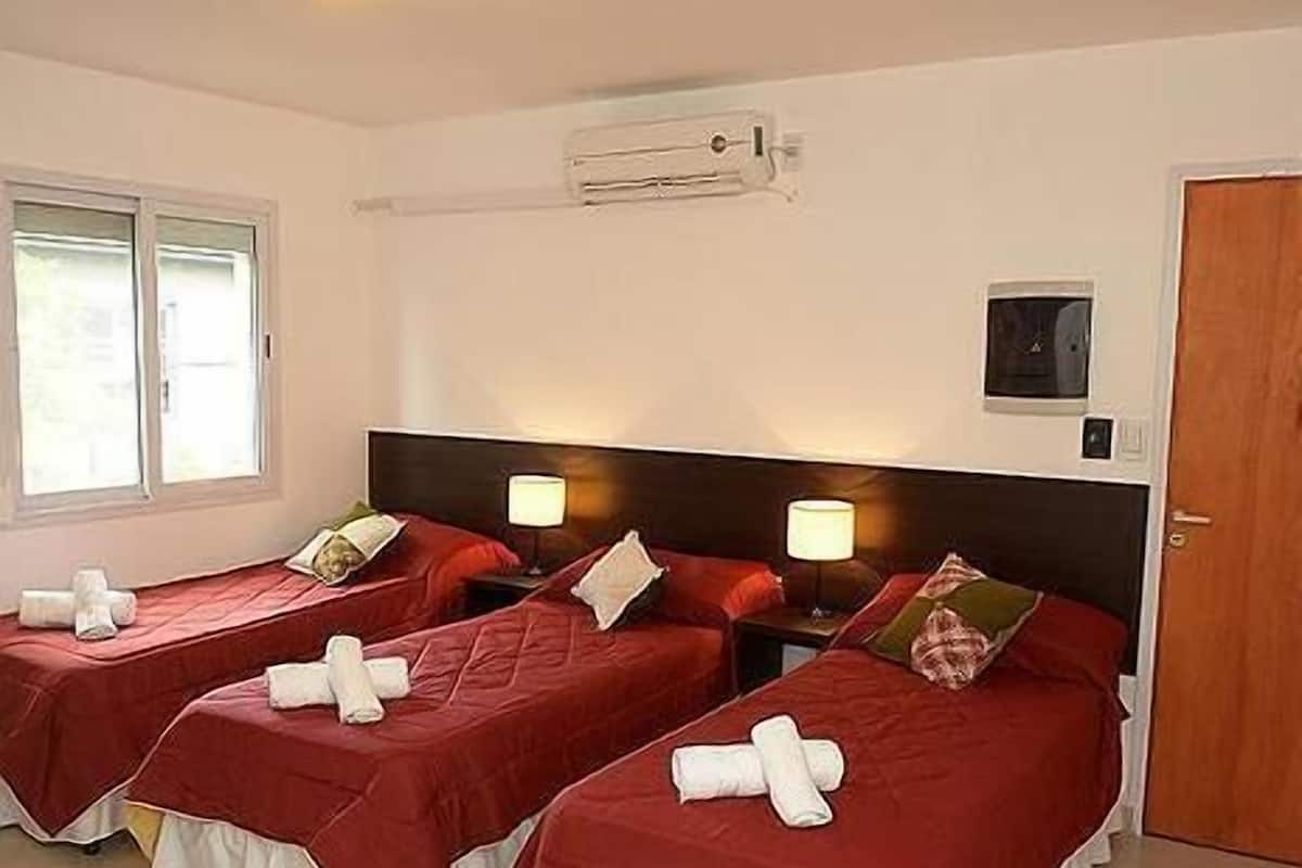 A hotel room with two single beds, each with a red bedspread and white pillows, a bedside table with a lamp, and a window with blinds.