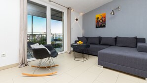 Ein modernes Wohnzimmer mit einem grauen Sofa, einem schwarzen Schaukelstuhl, einem kleinen runden Tisch mit Obst und einem großen Fenster mit Blick ins Freie.