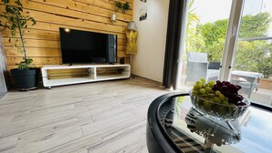 Ein modernes Wohnzimmer mit einer Holzverkleidung, einem Flachbildfernseher, einem Glastisch mit Obstschale und Blick ins Grüne.