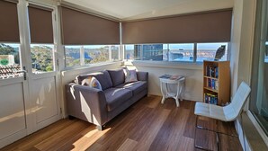 Ein modernes Wohnzimmer mit einer Couch, einem kleinen Tisch und einem Bücherregal. Große Fenster bieten Blick auf die Stadtlandschaft.