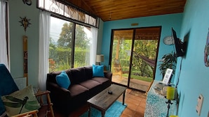 A cozy living room with a brown sofa, blue cushions, a wooden coffee table, and a view of lush greenery outside. - Romantic Hidden Jungle Gem for Two, Spectacular Views, Spa, A/C & Private Pool (El Castillo)