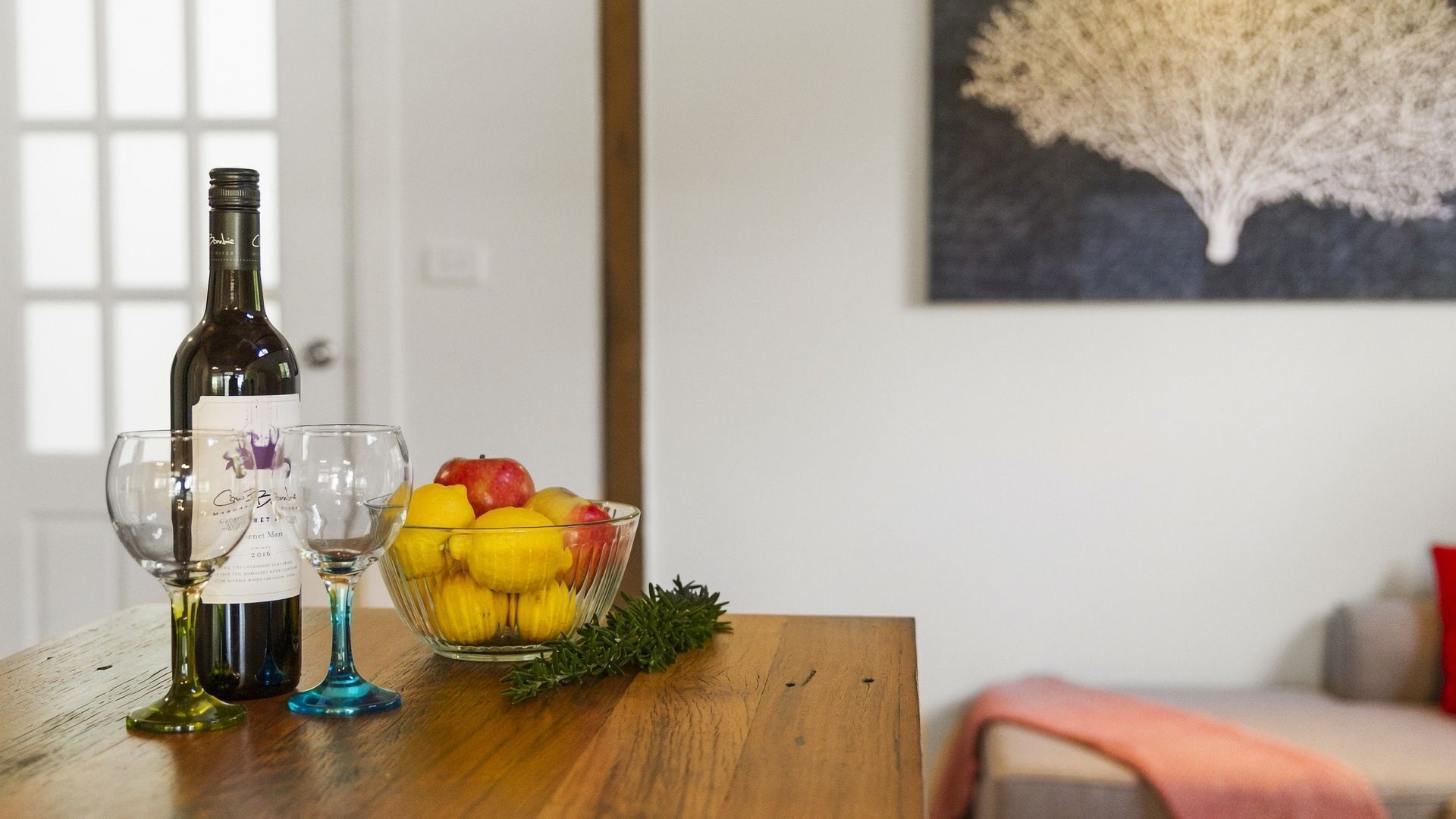 A wooden table with a wine bottle, two wine glasses, and a bowl of fruit.