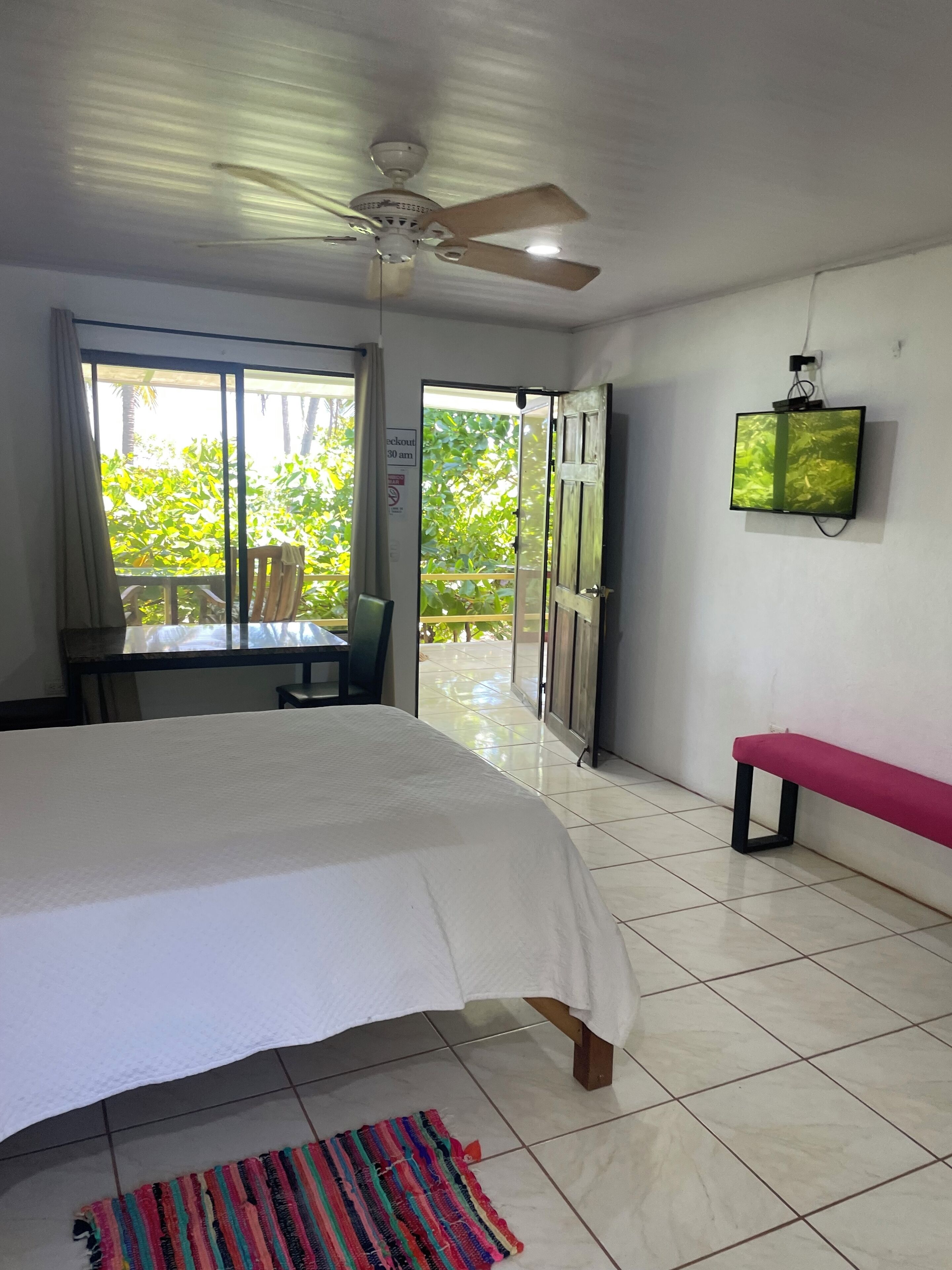 Habitación de hotel con cama, una banca, televisor, ventilador de techo y vista a zonas verdes.