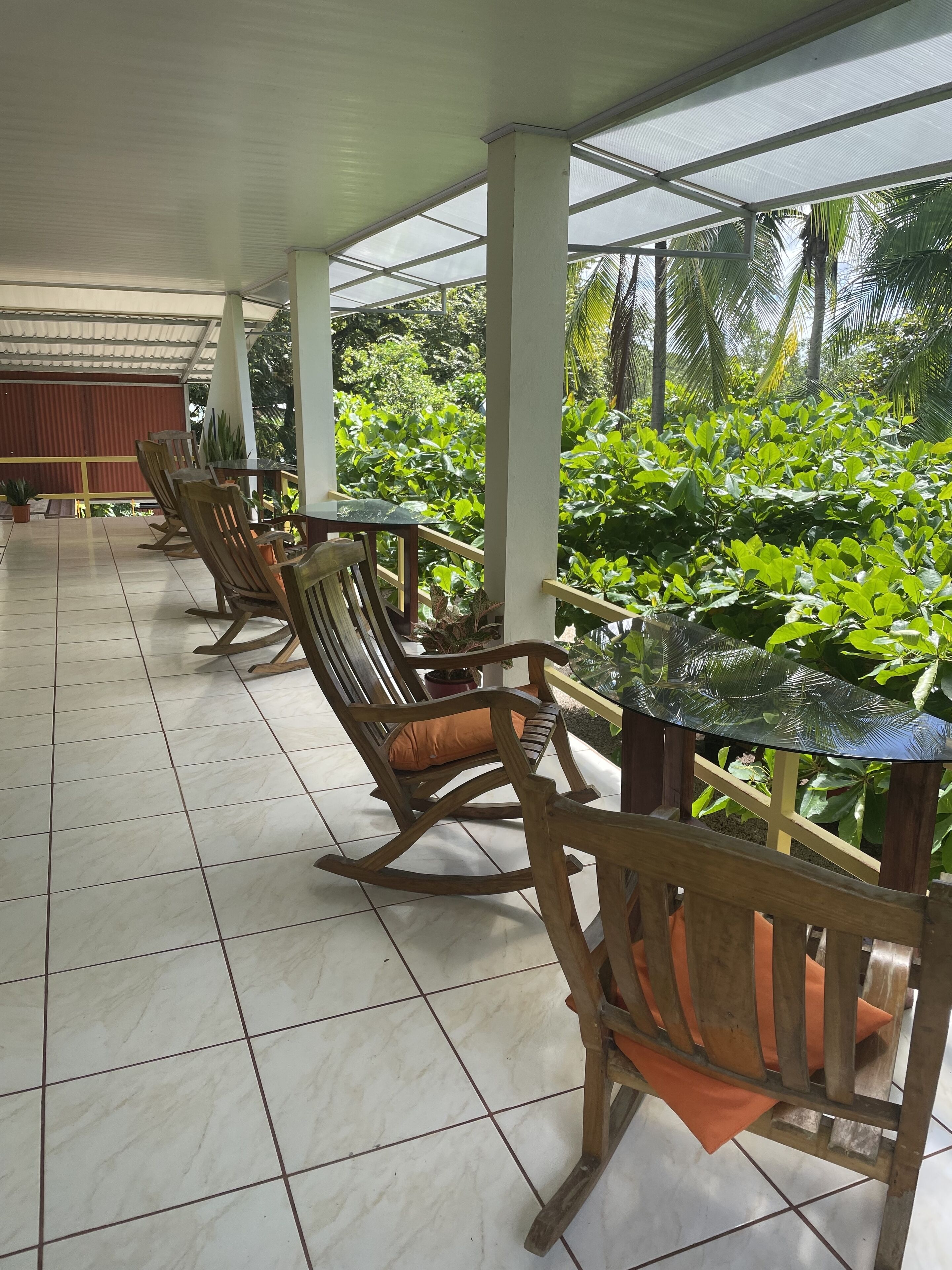 A covered outdoor seating area with wooden rocking chairs and a glass table, surrounded by lush greenery.