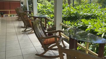 A covered outdoor seating area with wooden rocking chairs and a glass table, surrounded by lush greenery.