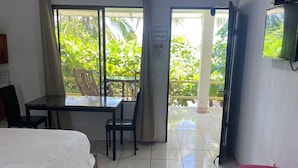 A hotel room with a bed, a table, and a bench. There is a ceiling fan, a TV mounted on the wall, and a window with curtains.