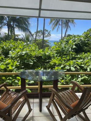 Une table en verre, entourée de chaises en bois, dans un espace extérieur abrité offrant une vue sur l’océan et les palmiers.