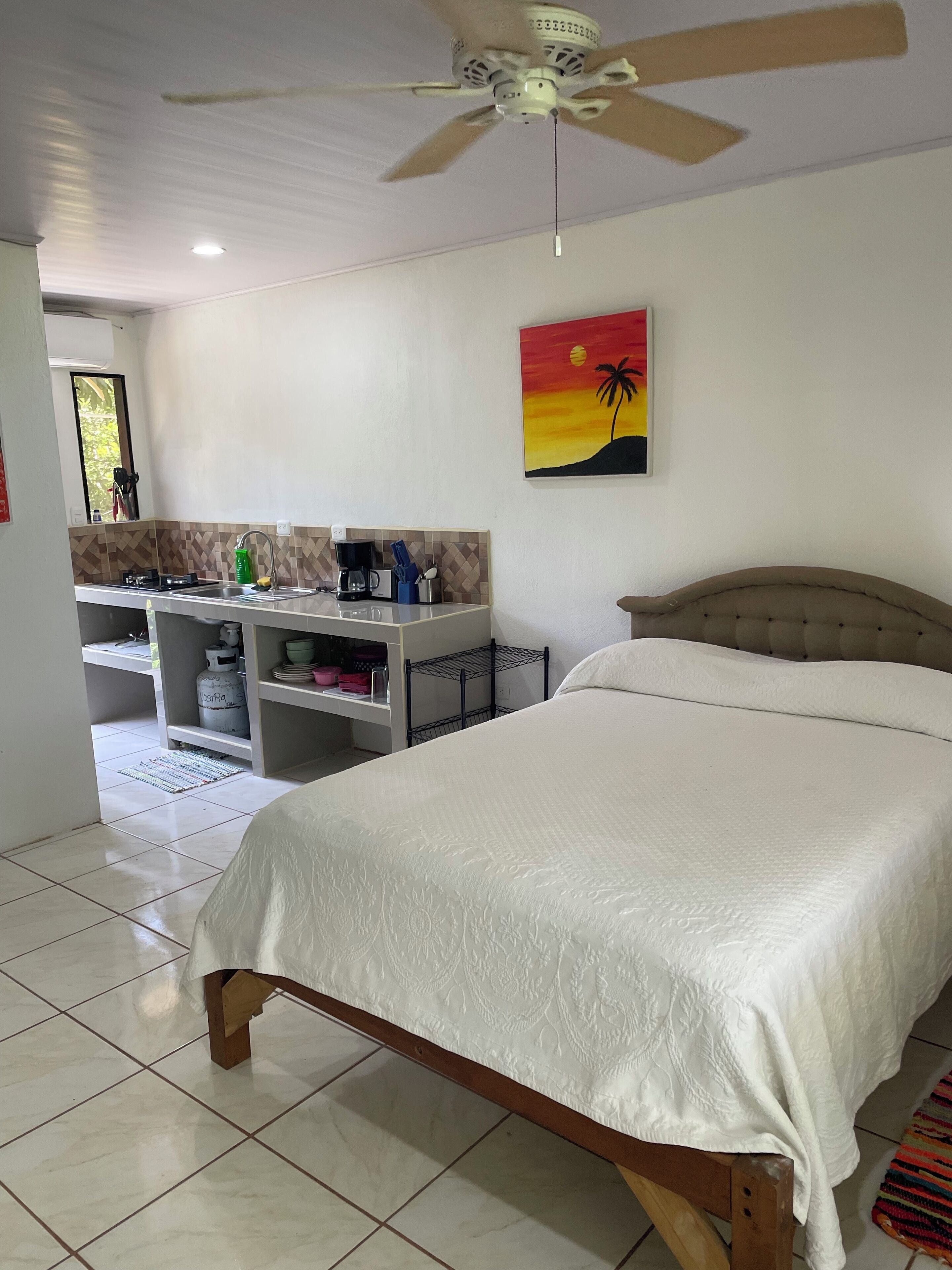 A bedroom with a bed, a ceiling fan, a kitchenette, and a painting of a sunset.