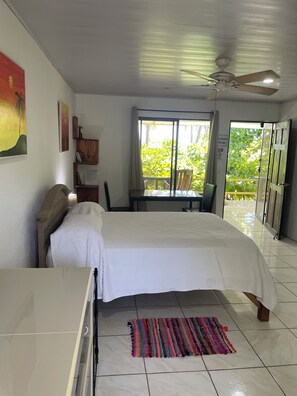 Une chambre à coucher comprenant un lit, un bureau, une chaise, un ventilateur de plafond, une fenêtre donnant sur un espace verdoyant et une porte menant à l’extérieur.