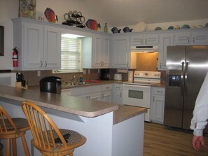 A kitchen with white cabinets, a stainless steel refrigerator, a stove, and a sink. There are two wooden chairs and a window with blinds. - Quiet Bay House Retreat With Easy Access, Great View. (Port O'Conner)