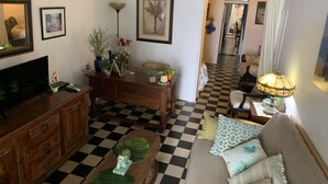 Un salon avec un sol à carreaux, une table basse en bois, un canapé agrémenté de coussins à motifs, un téléviseur, un bureau avec une lampe et des tableaux encadrés accrochés au mur.
