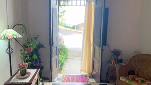 Une porte ouverte donnant sur la rue, avec un sol à carreaux, une chaise en osier, une lampe et une plante en pot à l’intérieur.