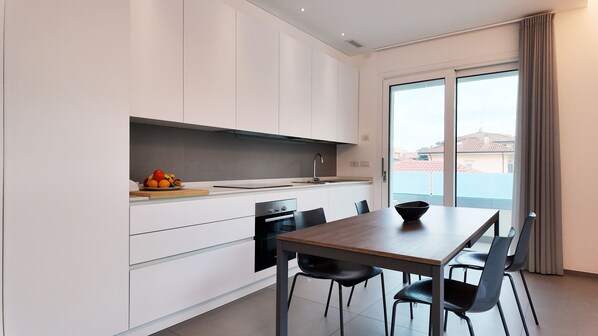 A modern kitchen with white cabinets, a wooden dining table, and a view of buildings through large windows. - New apartment in Viserba di Rimini with large garage, close to the sea! (Rimini)