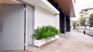 Moderne gevelingang met een glazen schuifdeur, een plantenbak met groene beplanting, en een betegeld voetpad dat uitkomt op een straat met andere gebouwen.