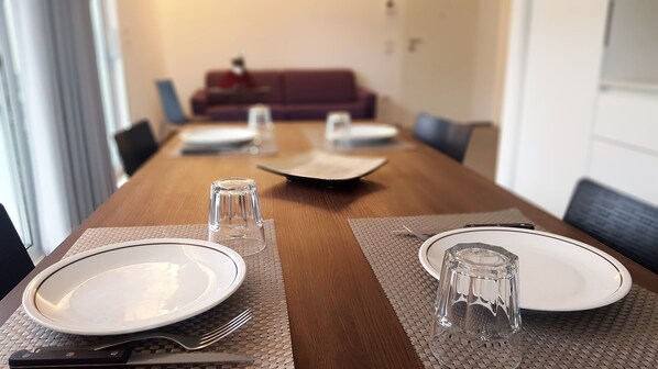 A dining table set with plates, cutlery, and glasses, with a sofa and chairs in the background. - New apartment in Viserba di Rimini with large garage, close to the sea! (Rimini)