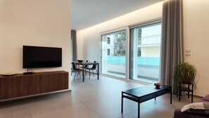 Een moderne woonkamer met een flatscreen televisie, een eethoek met een tafel en stoelen, en een grote schuifpui die leidt naar een buitenbalkon.