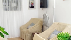 Ein Zimmer mit Esstisch, zwei beigen Sesseln, einem weißen Ventilator, einer Topfpflanze und einem Fernseher an der Wand.