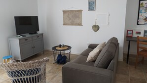 Un salon avec un canapé gris, un fauteuil en osier, une petite table ronde surmontée d’un set de table en fibres naturelles, un téléviseur posé sur un meuble gris et un mur orné d’une carte et de tableaux encadrés.