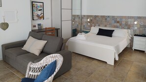 Une chambre d’hôtel moderne avec un canapé gris, un fauteuil en osier, un lit soigneusement fait et un petit bureau avec une chaise.
