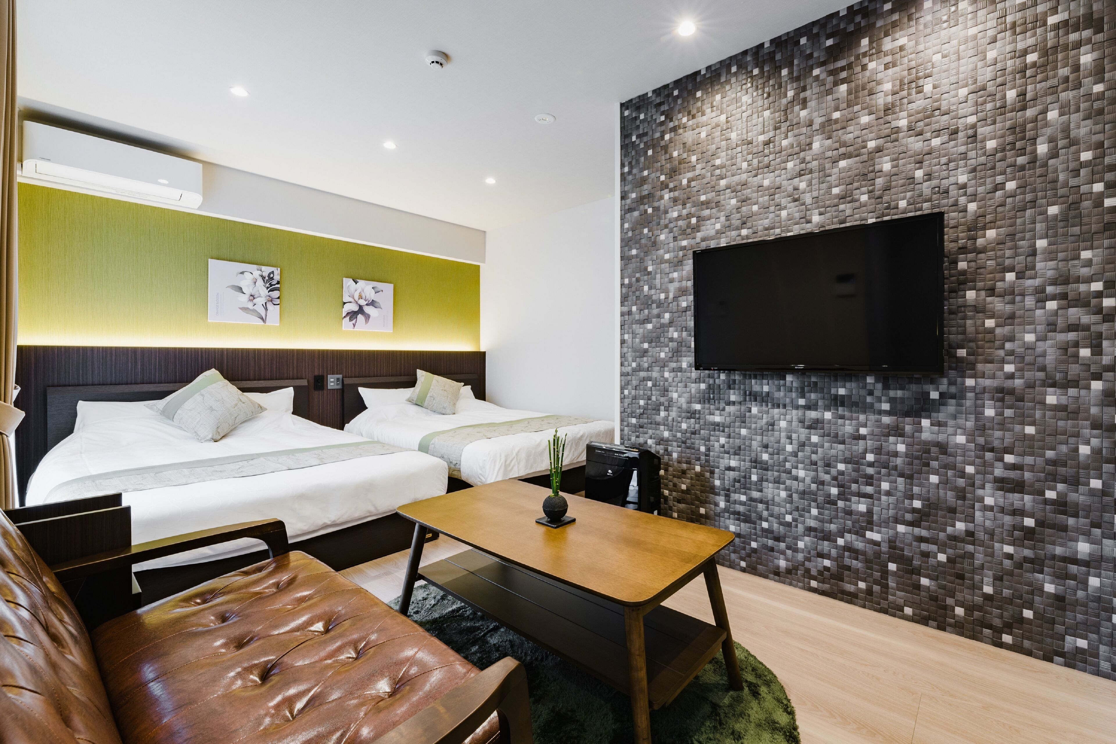 A modern hotel room with two beds, a flat-screen TV mounted on a mosaic wall, a wooden coffee table, and a leather sofa.