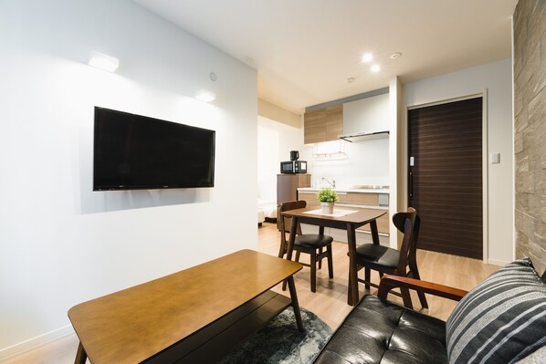 A modern living room with a flat-screen TV mounted on the wall, a wooden dining table with chairs, and a leather sofa.