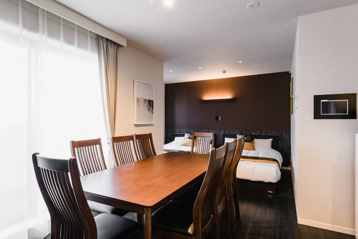 A hotel room with a wooden dining table, six chairs, a bed with a headboard, and a wall-mounted TV.